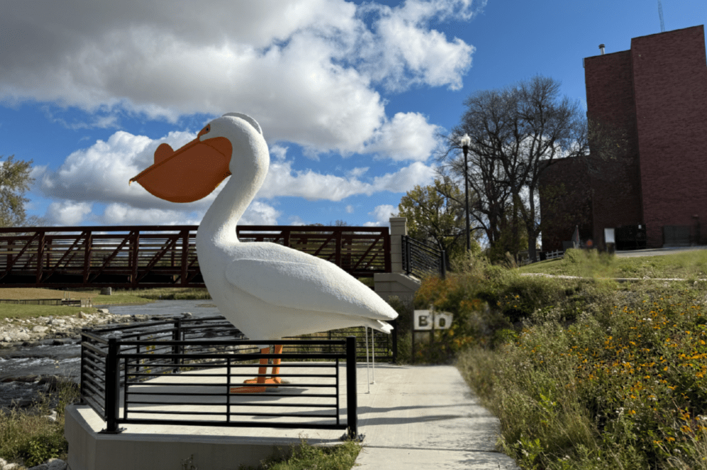 picture of a pelican--white body, orange beak. standing on a type of fenced overlook. blue sky and a bridge in the background