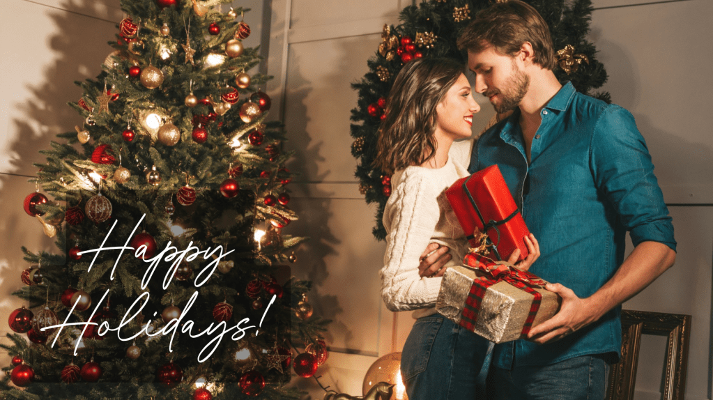 couple standing near christmas tree holding presents. she's wearing a white sweater, he's wearing a teal dress shirt both in jeans